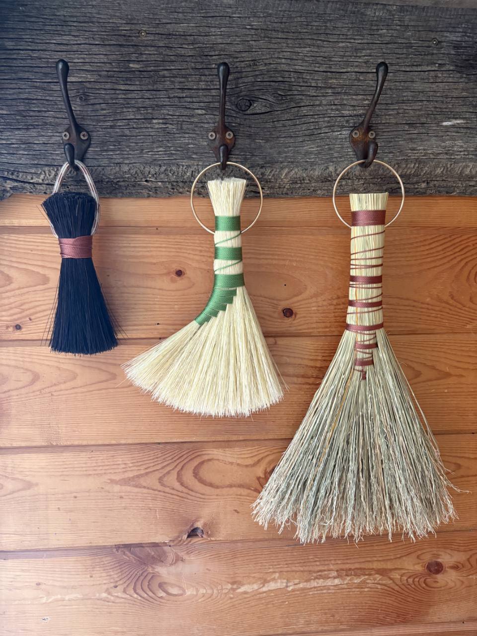 Three handmade Swale and Spry brooms hanging from hooks on a rustic wood wall.
